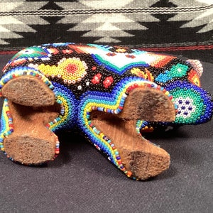 May include: A colorful beaded bear figurine with a black, blue, red, yellow, and green color scheme. The bear is standing on four wooden legs.