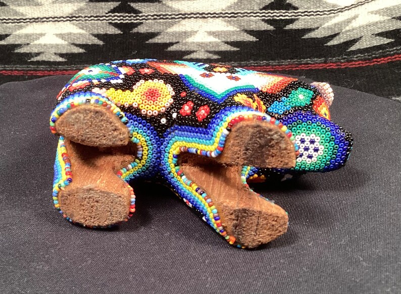 May include: A colorful beaded bear figurine with a black, blue, red, yellow, and green color scheme. The bear is standing on four wooden legs.