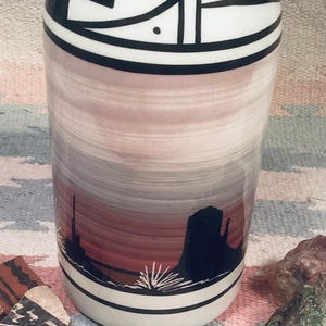May include: A ceramic vase with a southwestern design. The vase is white with black and red accents. It features a sunset scene with a mountain silhouette and a desert landscape.
