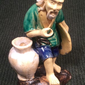 May include: A ceramic figurine of a man with a long white beard and mustache, wearing a green and blue robe, holding a pipe and sitting on a rock with a large jar beside him.