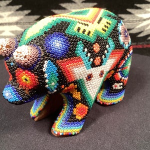 May include: A colorful beaded bear figurine with a geometric pattern. The bear is made of small, round beads in shades of blue, green, red, yellow, and white. The bear is standing on a black surface.