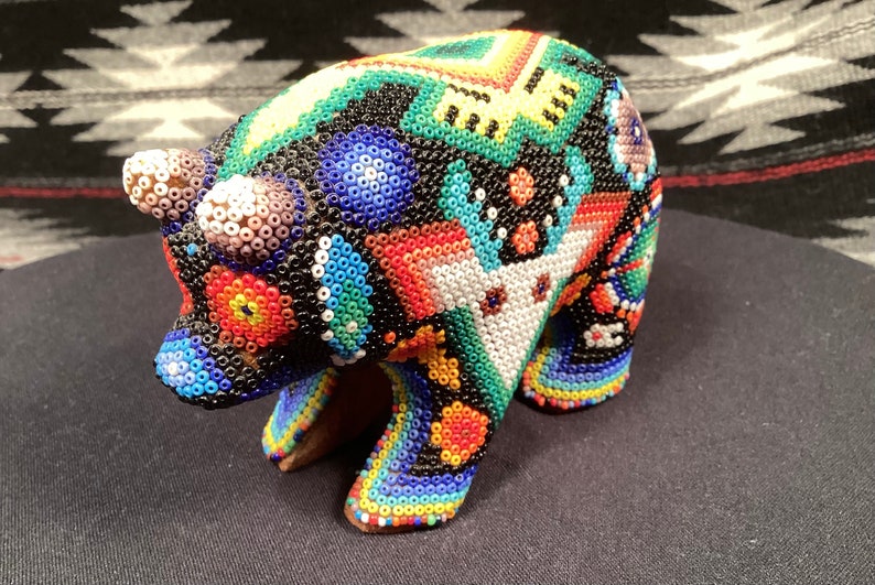 May include: A colorful beaded bear figurine with a geometric pattern. The bear is made of small, round beads in shades of blue, green, red, yellow, and white. The bear is standing on a black surface.