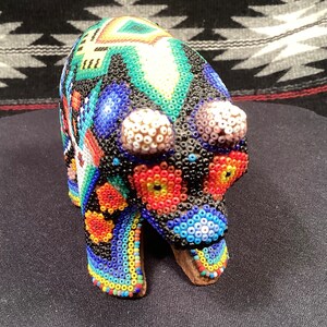 May include: A beaded bear figurine with a colorful geometric pattern. The bear is black with blue, green, red, yellow, and white beads.