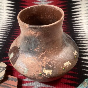 May include: A handcrafted, brown ceramic vase with a wide base and narrow neck. The vase features painted images of a building and animals. The exterior has a textured, aged appearance, with visible cracks and variations in color.