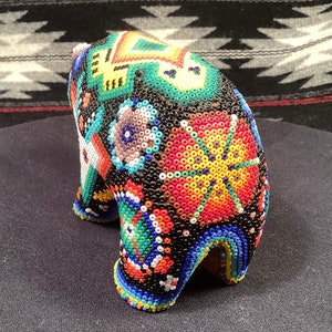 May include: A small, beaded bear figurine with a colorful geometric pattern. The bear is black with bright colors like red, yellow, green, blue, and white.