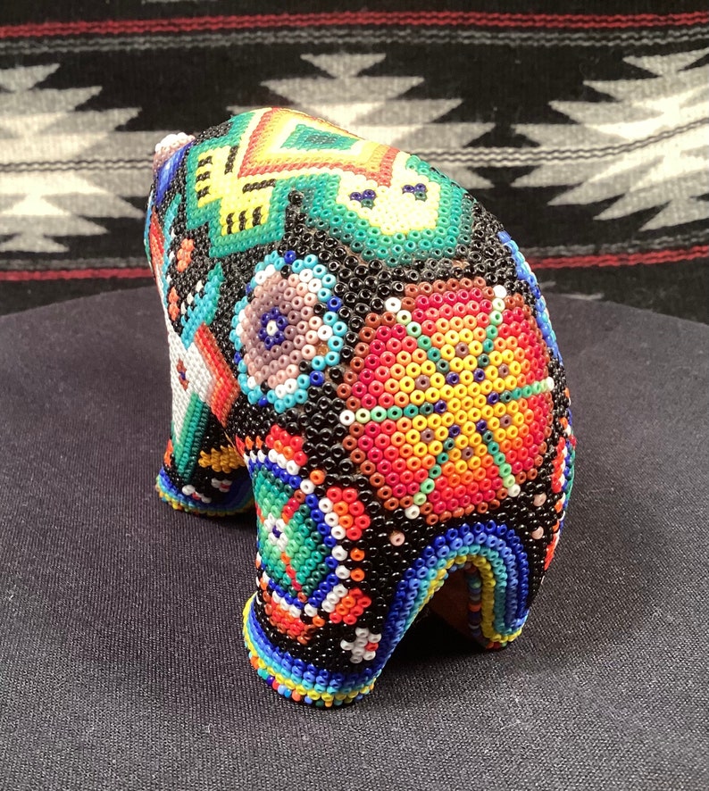 May include: A small, beaded bear figurine with a colorful geometric pattern. The bear is black with bright colors like red, yellow, green, blue, and white.