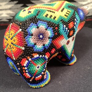 May include: A small, beaded bear figurine with a colorful, geometric pattern. The bear is made of black beads with red, orange, yellow, green, blue, and white beads forming the pattern.