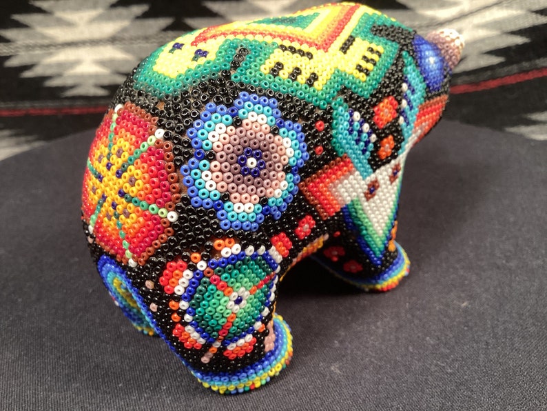 May include: A small, beaded bear figurine with a colorful, geometric pattern. The bear is made of black beads with red, orange, yellow, green, blue, and white beads forming the pattern.