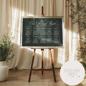 May include: A wedding seating plan displayed on a wooden easel. The chart features a dark green forest scene and white text, including "Our Favorite People" and "Your Seat Awaits." Names are listed under numbers.
