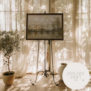 May include: A rectangular wedding seating chart sign with a dark frame, featuring the text "Our Favorite People" and "Your Seat Awaits" above ten numbered columns, displayed on a tall, thin, black metal tripod easel stand.