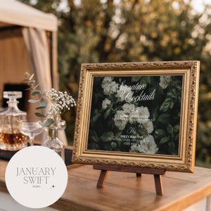 May include: A rectangular gold ornate frame holding a sign with white floral imagery and the text Signature Cocktails, Her Classic Margarita, and His Dirty Martini. The frame is displayed on a small wooden easel stand on a wooden surface.