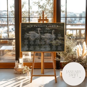 May include: A framed seating plan on a wooden easel, with the text "OUR FAVORITE PEOPLE" and "PLEASE FIND YOUR SEAT". The chart features a swan illustration and guest names. The easel is positioned in front of a large window.