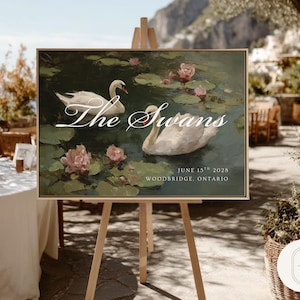 May include: A framed sign on a wooden easel displays a painting of two white swans in a pond with pink water lilies. The text "The Swans" is written in elegant script above the date and location.