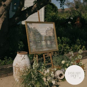 May include: A rectangular wedding seating chart sign with a gold frame, displayed on a wooden easel. The sign features a vintage-style landscape painting background with the text Our Favorite People and Your Seat Awaits, listing guest names by table.