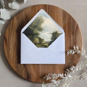 May include: A white envelope with a landscape painting inside, resting on a round wooden tray. The painting depicts trees, water, and a cloudy sky. Decorative white sprigs are placed near the tray.
