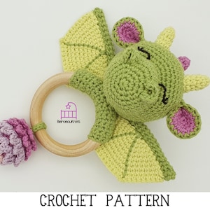 May include: Green crocheted dragon rattle with pink ears and a wooden ring. The dragon has a smiling face and a pink flower attached to the ring.