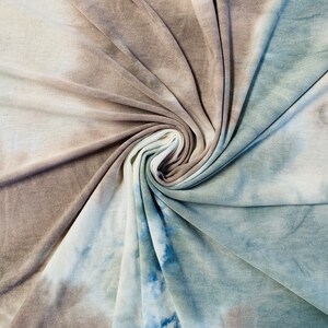 Könnte beinhalten: Ein weicher, dehnbare Stoff mit einem Batik-Muster in Braun-, Weiß- und Blautönen. Der Stoff ist gefaltet und verdreht, wodurch ein wirbelnder Effekt entsteht.