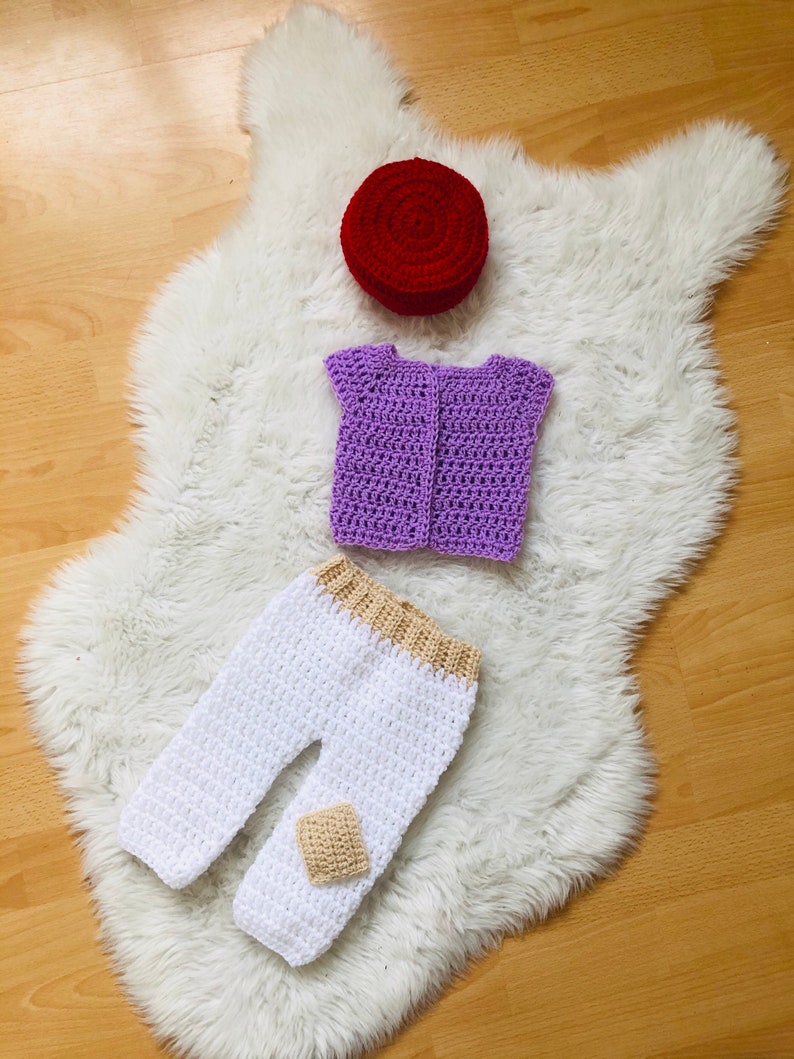 May include: A crocheted outfit for a baby, including a purple vest, white pants with a tan patch, and a red hat.