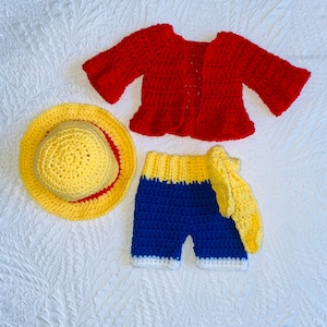 May include: A crocheted baby outfit in red, yellow, and blue. The outfit includes a hat, a cardigan, and shorts.