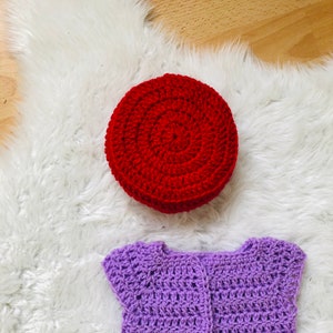 May include: A purple crocheted baby cardigan with short sleeves and a red crocheted hat.