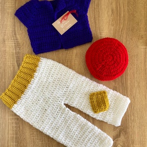 May include: A crocheted baby outfit in blue, white, and yellow with a red crocheted hat. The outfit includes a vest, pants, and a hat.