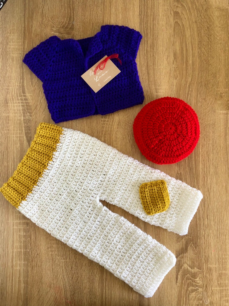 May include: A crocheted baby outfit in blue, white, and yellow with a red crocheted hat. The outfit includes a vest, pants, and a hat.