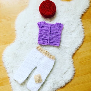 May include: A crocheted outfit for a baby, including a purple vest, white pants with a tan trim, and a red hat.