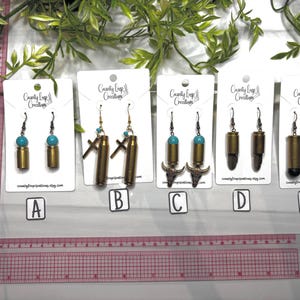May include: Five pairs of handmade earrings. Each pair features a brass bullet casing. Some have turquoise beads, crosses, bull heads, or black accents. The earrings are displayed on white cards with the text "County Loop Creations".