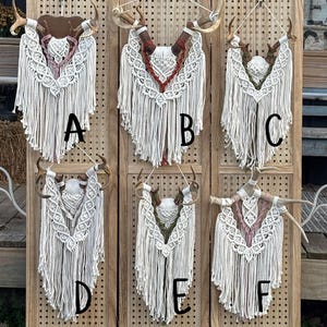 May include: A collection of six macrame wall hangings, each featuring deer antlers and intricate woven designs. The macrame is primarily white, with accents of pink, red, and green. The antlers are various shades of brown and beige, mounted on a wooden background.