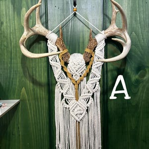 May include: A decorative wall hanging featuring deer antlers and a macrame design. The antlers are a light brown color, and the macrame is white and tan. The wall hanging is displayed against a green wooden background.