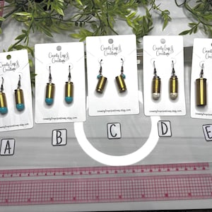 May include: A collection of handmade earrings displayed on white cards. The earrings feature brass bullet casings with various stone or bead accents, including turquoise and white stone. The earrings are presented against a backdrop of greenery and a pink ruler.