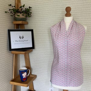 May include: A pale pink scarf with a geometric design is displayed on a mannequin. The pattern features small, repeating shapes in pink, blue, and white. A framed sign and a mug are on a wooden stand.
