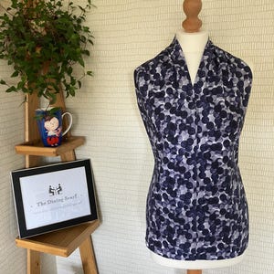 May include: A sleeveless top draped on a mannequin, featuring a repeating pattern of dark navy blue and light grey circles. The fabric has a soft, fluid appearance, and the garment is styled with a V-neckline that falls straight down the torso.