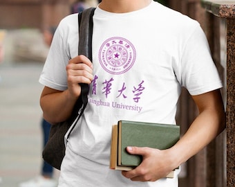 May include: A young man wearing a white t-shirt with a purple Tsinghua University logo and Chinese text. He is smiling and holding a stack of books. He is standing in front of a building with other students in the background.