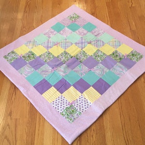 May include: A patchwork quilt with a purple, yellow, and green color scheme. The quilt is made of various fabrics with different patterns, including floral, plaid, and polka dots. The quilt is edged with a light purple fabric.