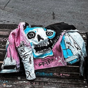 K&ouml;nnte beinhalten: Eine rosa und blaue Jeansjacke mit einem auf der Vorderseite gemalten Totenkopf. Der Totenkopf ist in Blau, Wei&szlig; und Schwarz bemalt. Die Jacke hat die Worte "Never Say The" auf dem linken &Auml;rmel.