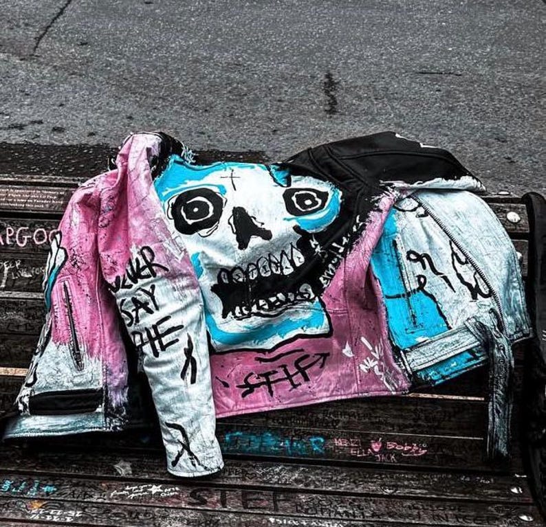 K&ouml;nnte beinhalten: Eine rosa und blaue Jeansjacke mit einem auf der Vorderseite gemalten Totenkopf. Der Totenkopf ist in Blau, Wei&szlig; und Schwarz bemalt. Die Jacke hat die Worte "Never Say The" auf dem linken &Auml;rmel.