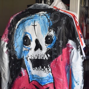 K&ouml;nnte beinhalten: Eine schwarze Lederjacke mit einem bemalten Totenkopf-Design. Der Totenkopf ist in Blau, Wei&szlig; und Schwarz mit einem Kreuz auf der Stirn bemalt. Die Jacke hat auch rosa und wei&szlig;e Farbschleier. Der Text "NEVER SAY DIE" ist auf der Jacke geschrieben.