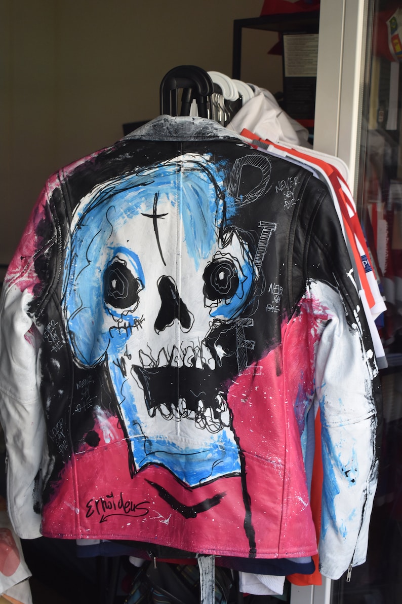K&ouml;nnte beinhalten: Eine schwarze Lederjacke mit einem bemalten Totenkopf-Design. Der Totenkopf ist in Blau, Wei&szlig; und Schwarz mit einem Kreuz auf der Stirn bemalt. Die Jacke hat auch rosa und wei&szlig;e Farbschleier. Der Text "NEVER SAY DIE" ist auf der Jacke geschrieben.