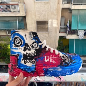 Könnte beinhalten: Ein High-Top-Sneaker mit handbemaltem Design, das eine markante schwarz-weiße Totenkopf-Grafik auf weißem Grund zeigt. Der Schuh ist mit kräftigen blauen und roten Farbakzenten versehen und trägt den weißen Schriftzug DIE.