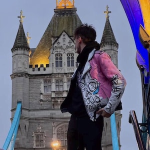 K&ouml;nnte beinhalten: Eine Person in einer rosa und wei&szlig; bemalten Lederjacke steht vor der Tower Bridge in London, England. Die Jacke hat ein Totenkopfdesign auf der Vorderseite.