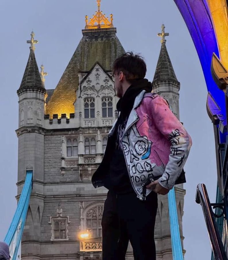 K&ouml;nnte beinhalten: Eine Person in einer rosa und wei&szlig; bemalten Lederjacke steht vor der Tower Bridge in London, England. Die Jacke hat ein Totenkopfdesign auf der Vorderseite.