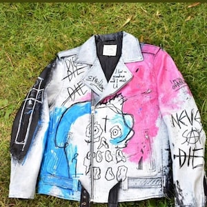 K&ouml;nnte beinhalten: Eine wei&szlig;e Lederjacke mit blauen und pinkfarbenen Farbflecken und einem Totenkopf-Motiv. Die Jacke hat einen Rei&szlig;verschluss und einen schwarzen G&uuml;rtel. Der Text "DIE" ist in schwarzer Farbe auf die Jacke geschrieben.