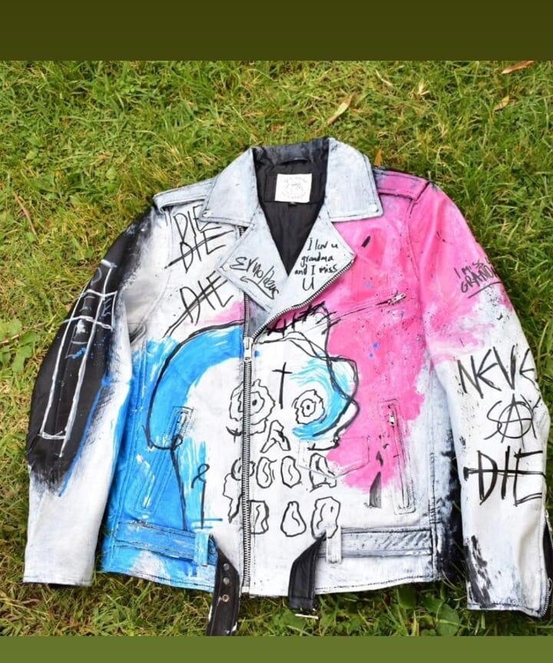 K&ouml;nnte beinhalten: Eine wei&szlig;e Lederjacke mit blauen und pinkfarbenen Farbflecken und einem Totenkopf-Motiv. Die Jacke hat einen Rei&szlig;verschluss und einen schwarzen G&uuml;rtel. Der Text "DIE" ist in schwarzer Farbe auf die Jacke geschrieben.