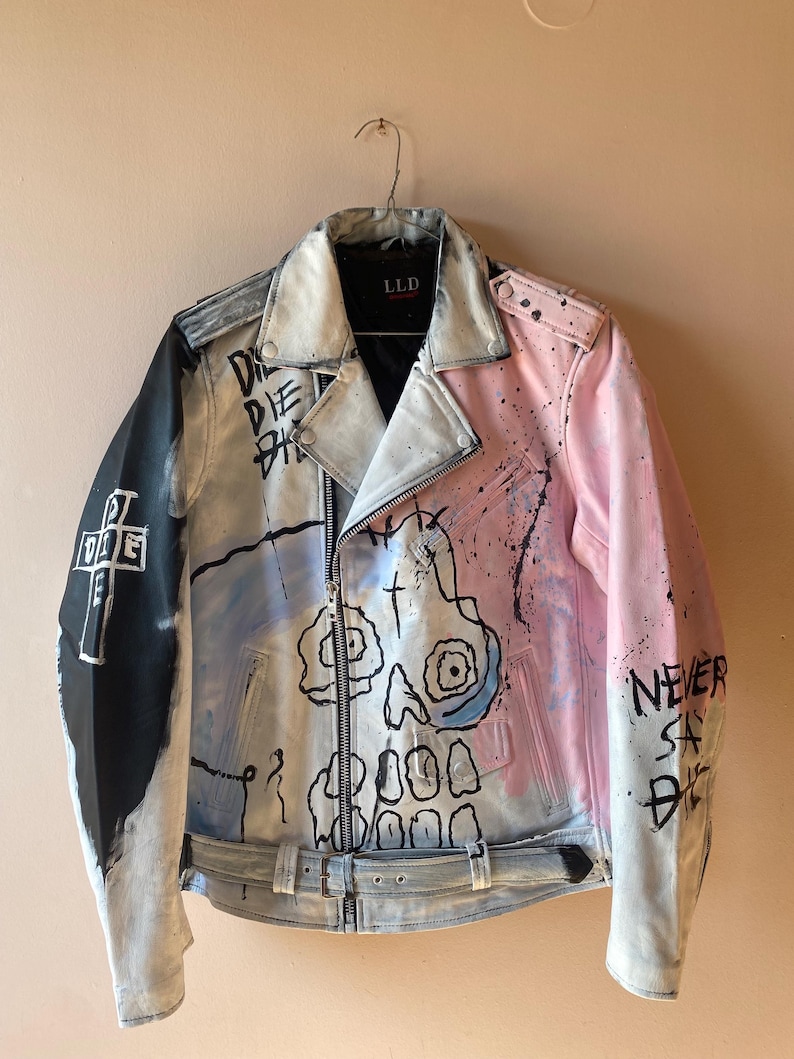 K&ouml;nnte beinhalten: Eine wei&szlig;e, rosa und blaue Lederjacke mit einem Totenkopf-Motiv und dem Text "DIE DIE DIE" und "NEVER SAY DIE", der darauf gemalt ist. Die Jacke hat einen Rei&szlig;verschluss und einen G&uuml;rtel an der Taille.