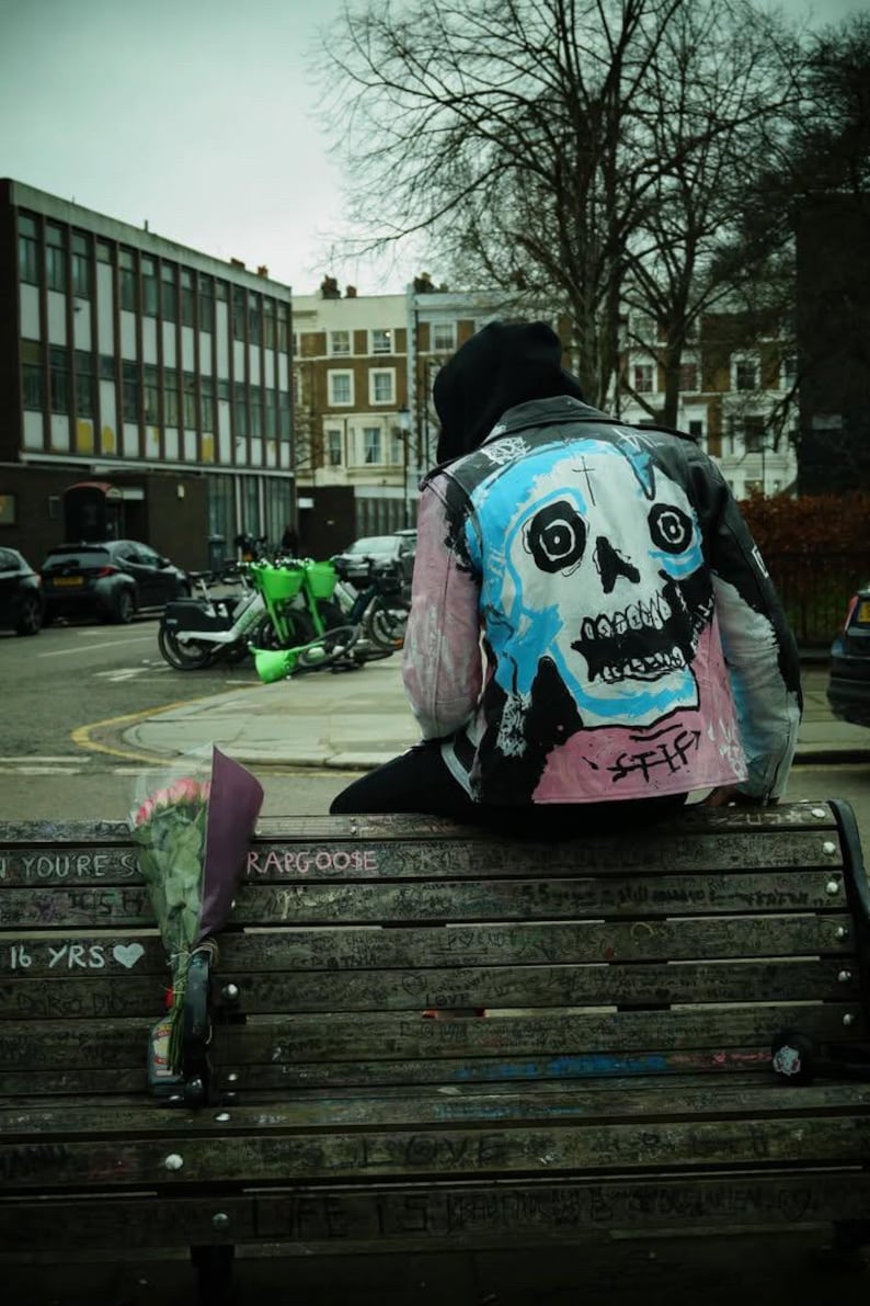 K&ouml;nnte beinhalten: Eine Person tr&auml;gt einen schwarzen Kapuzenpullover und eine rosafarbene und blaue Lederjacke mit einem bemalten Totenkopfdesign und sitzt auf einer Holzbank. Die Bank ist mit Graffiti bedeckt und hat die Worte "You're so 16 yrs" und "Ragoose" darauf geschrieben.