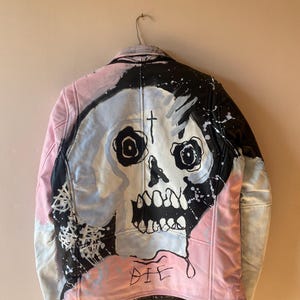 K&ouml;nnte beinhalten: Eine schwarze Lederjacke mit einem pinkfarbenen und blauen bemalten Sch&auml;delmotiv auf der R&uuml;ckseite. Der Sch&auml;del hat ein Kreuz auf der Stirn und das Wort "DIE" ist darunter geschrieben.