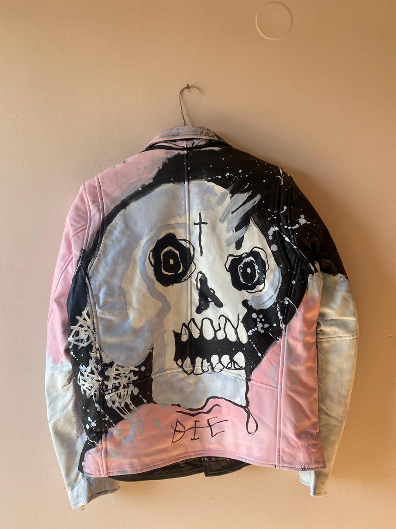K&ouml;nnte beinhalten: Eine schwarze Lederjacke mit einem pinkfarbenen und blauen bemalten Sch&auml;delmotiv auf der R&uuml;ckseite. Der Sch&auml;del hat ein Kreuz auf der Stirn und das Wort "DIE" ist darunter geschrieben.
