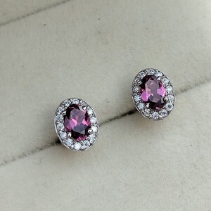 May include: A pair of silver earrings featuring oval-shaped purple gemstones encircled by a halo of smaller white gemstones.