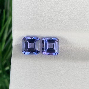May include: Two square-cut, light blue gemstones with a slight purple hue. The gems are displayed on a white surface, with green grass visible in the background. The gemstones are likely for jewelry.
